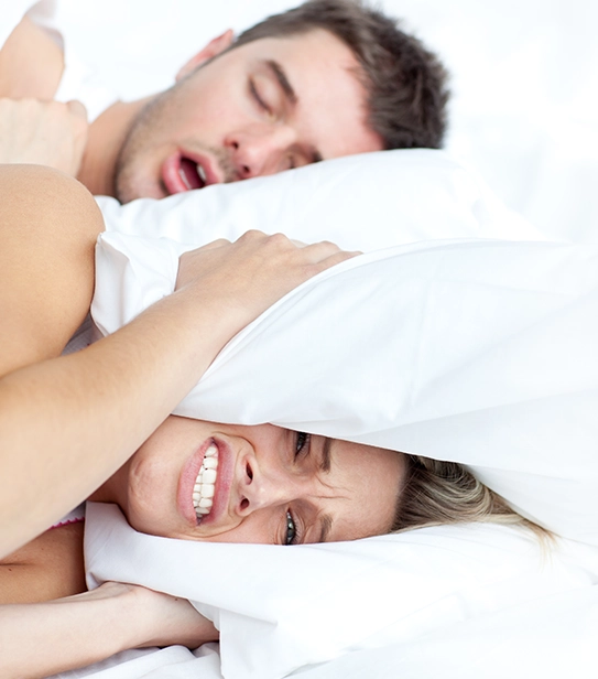 woman covering ears with pillow while partner snores loudly in bed
